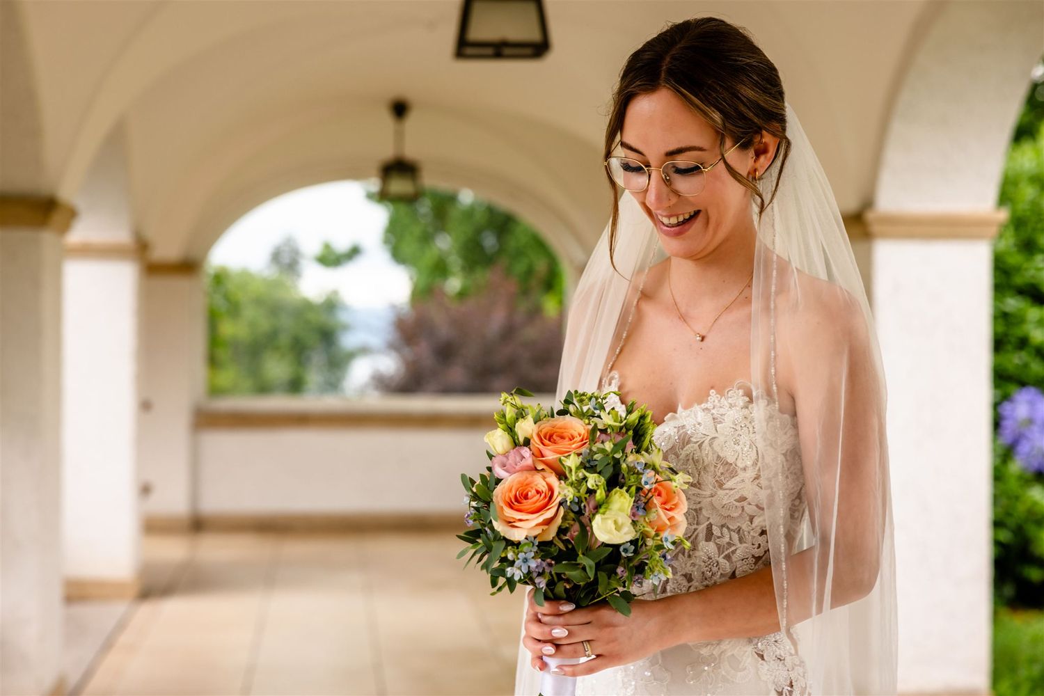 A bride in a white lace wedding dress holds a vibrant bouquet of orange and pink flowers in an outdoor setting.