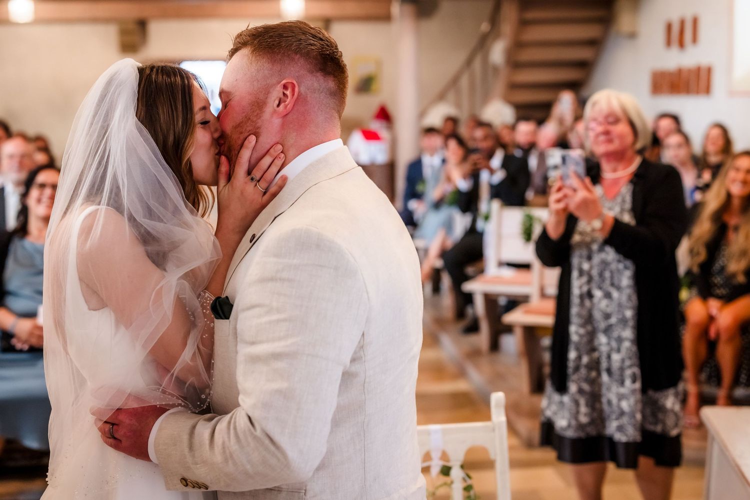 Preise für Hochzeitsfotografen Braut und Bräutigam teilen ihren ersten Kuss bei der Hochzeitszeremonie, während die Gäste in einer sonnenbes