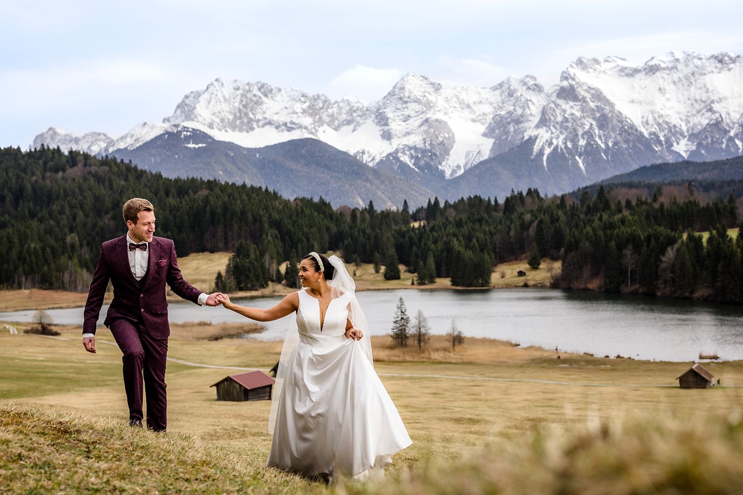 A couple in wedding attire dances together against a backdrop of snow-capped mountains and a serene lake.