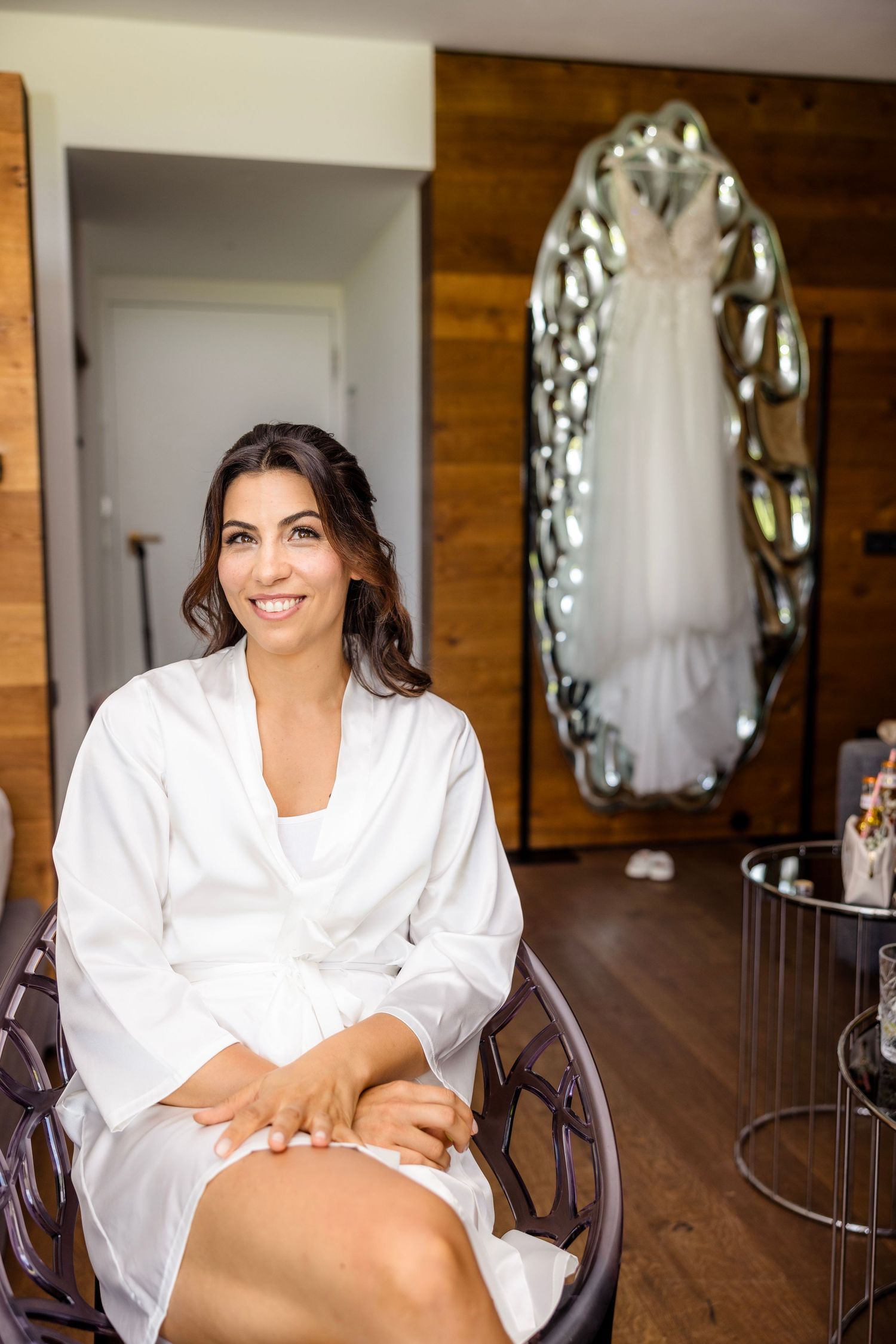 A woman in a white robe sits in a chair while a wedding dress hangs on display in an ornate mirror behind her.