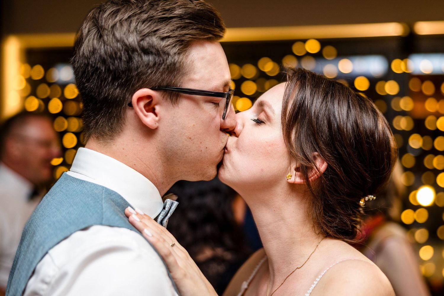 A romantic moment between a couple kissing at an evening celebration with glowing lights in the background.