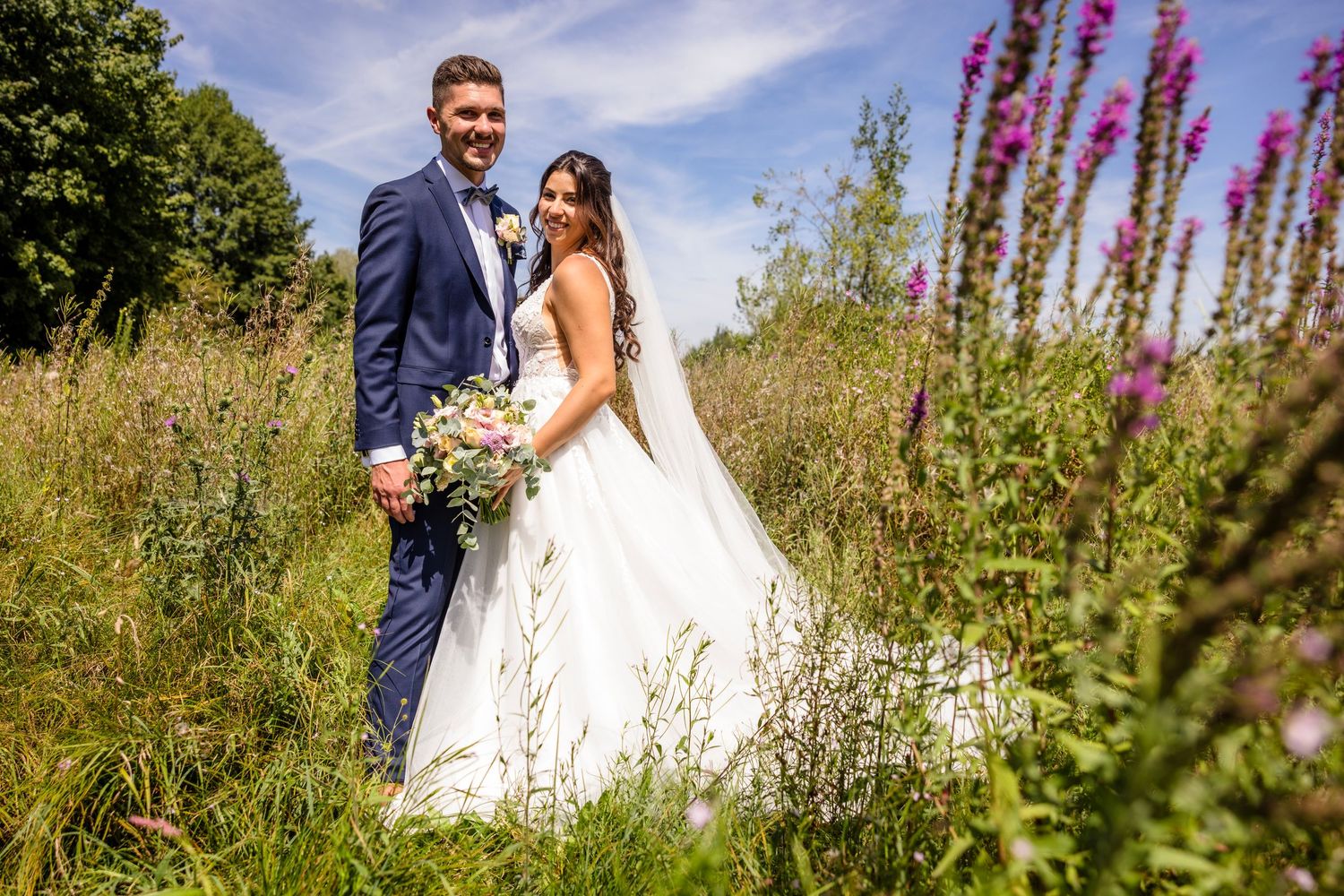 A couple shares a romantic moment among tall purple wildflowers in a summer meadow on their wedding day.