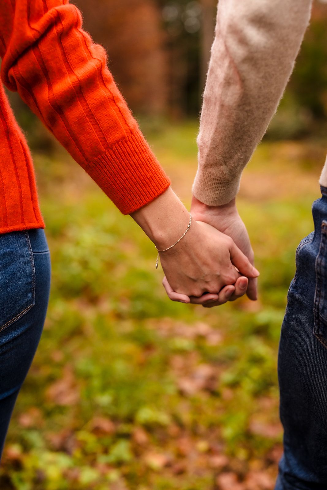 A couple holding hands outdoors wearing jeans and sweaters, with one person in a bright orange knit sweater.