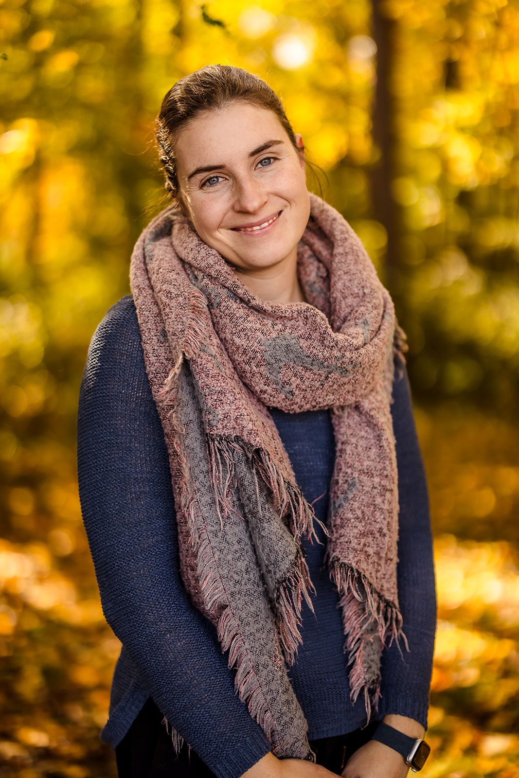 Person wearing navy sweater and pink scarf smiling in autumn forest setting.