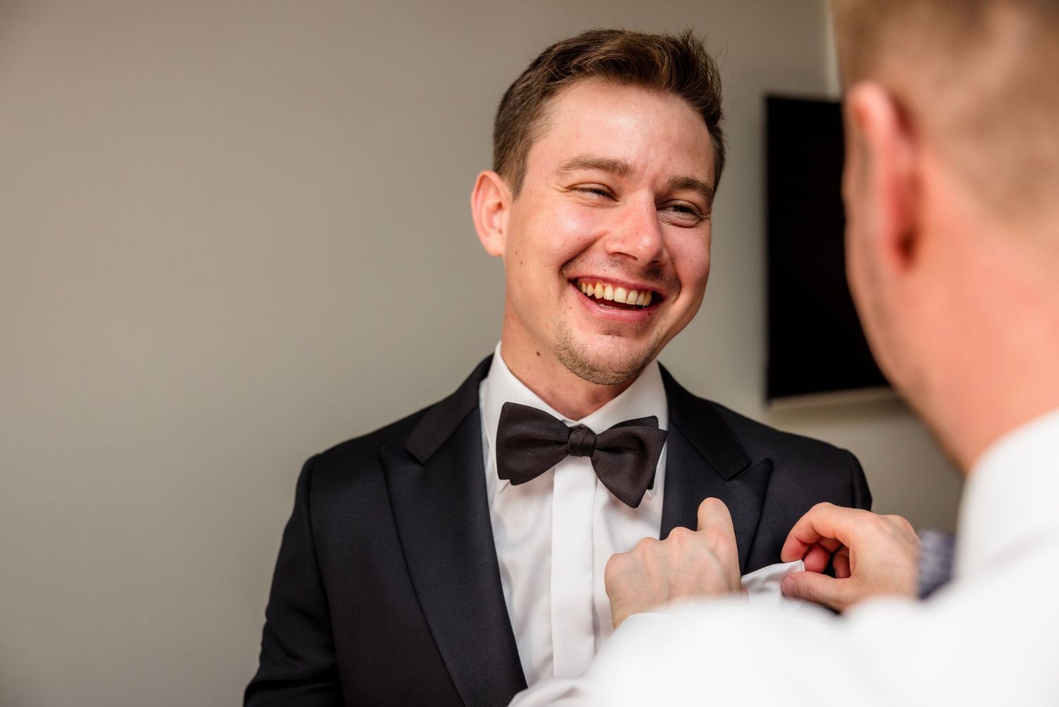 A laughing groom in a black tuxedo and bow tie gets his boutonniere pinned by a groomsman before his wedding.