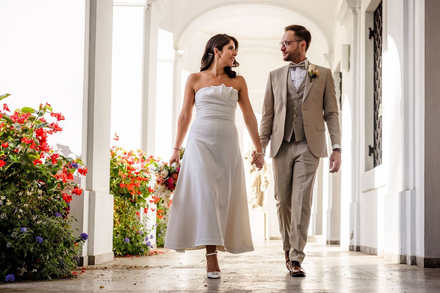 Bride and groom walking hand in hand through a sunlit colonnade adorned with colorful flowers on their wedding day.