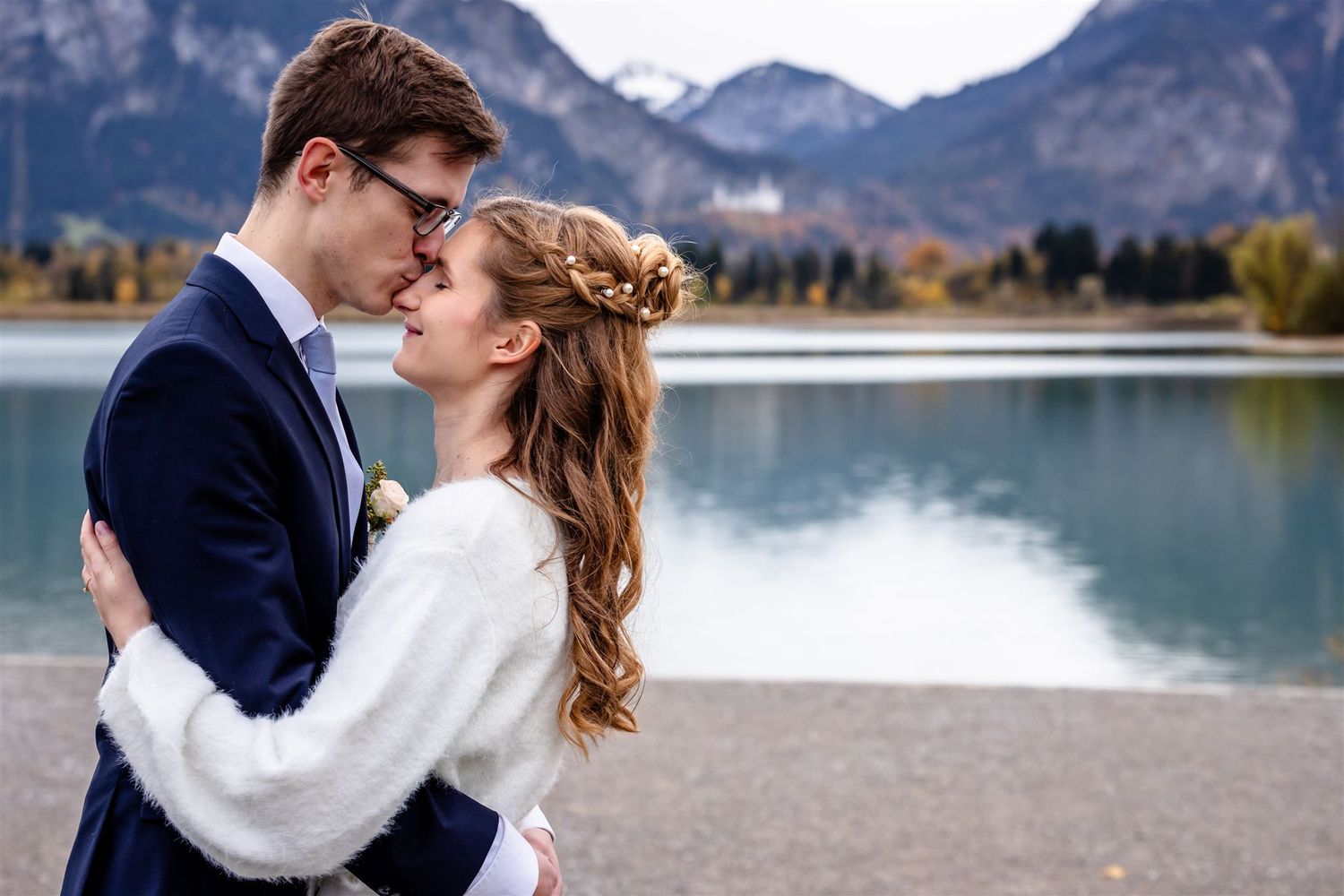 Romantic couple embraces and kisses by a scenic mountain lake with snow-capped peaks in the background.