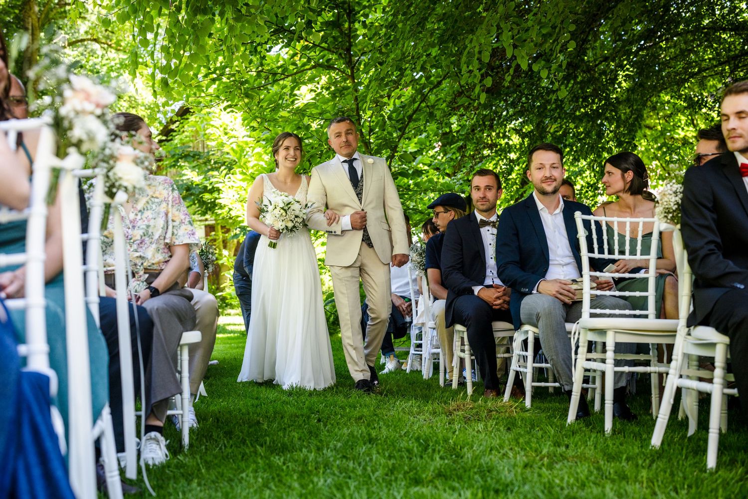 A couple walks down the outdoor garden aisle during a summer wedding ceremony with white chairs and lush greenery. Hofgut Sickertshofen