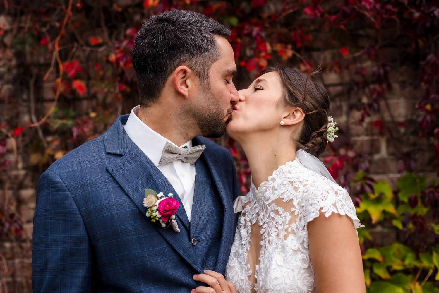 A bride and groom share a romantic kiss on their wedding day in front of a red ivy-covered brick wall.