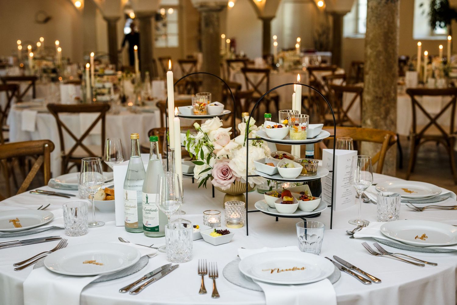 Elegant wedding reception tables set with white linens, floral centerpieces, and rustic wooden chairs in a warm-lit venue. Hofgut Sickertsho