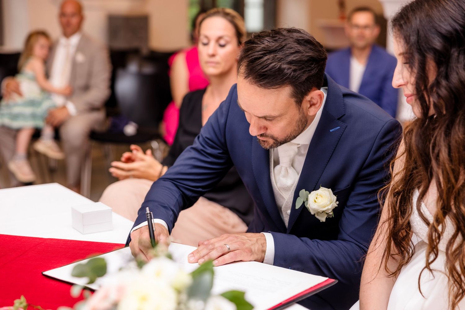 Groom in blue suit signing wedding register at ceremony table with bride and guests in background.