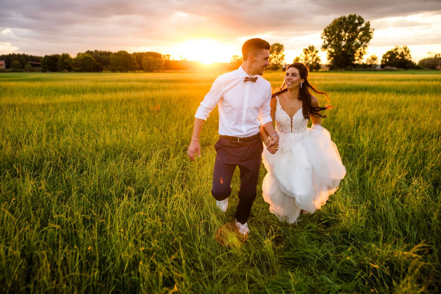 Bride and groom running joyfully through a lush green field at golden sunset on their wedding day.