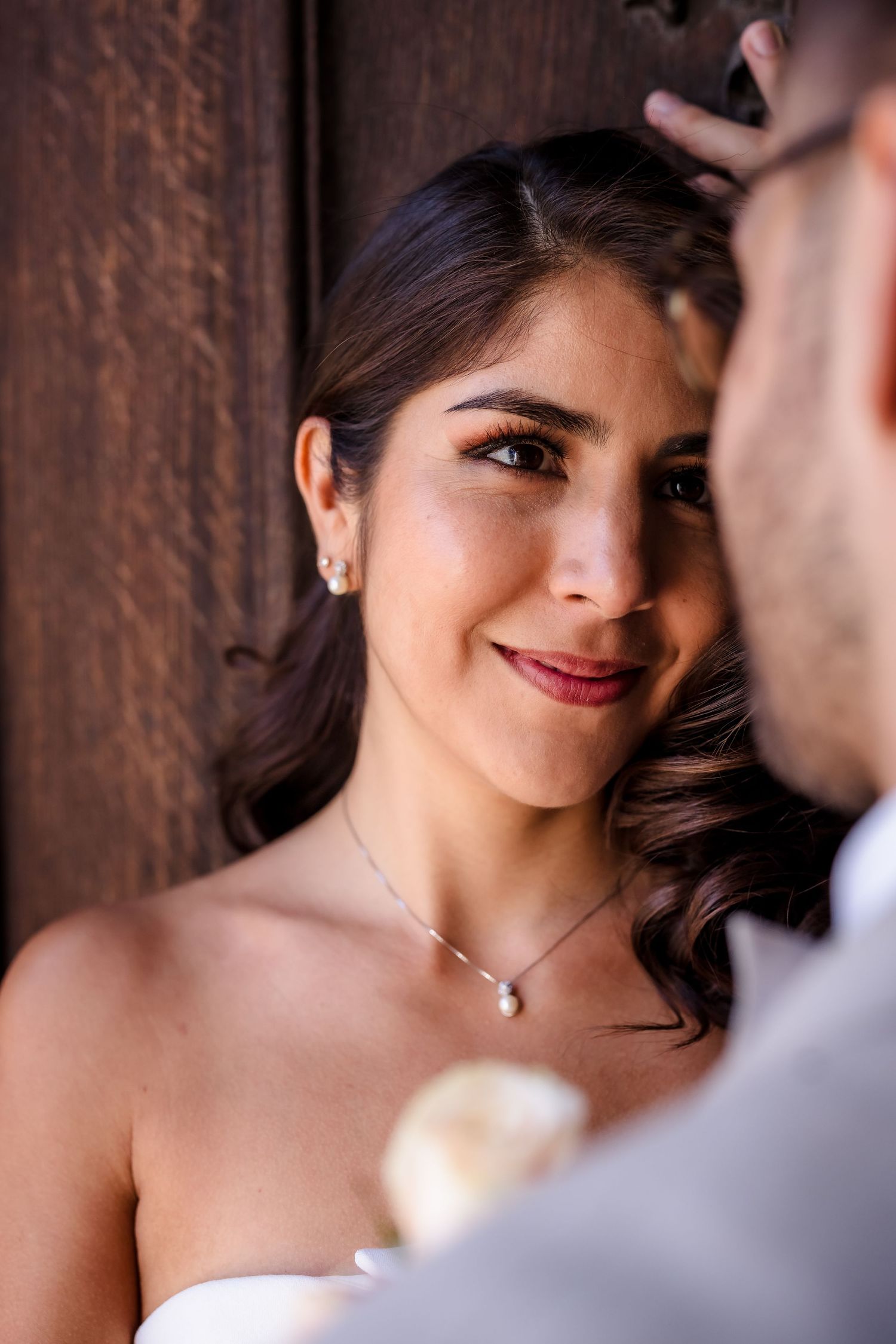 Smiling bride in white dress with pearl necklace gazes at groom against rustic wooden door backdrop.