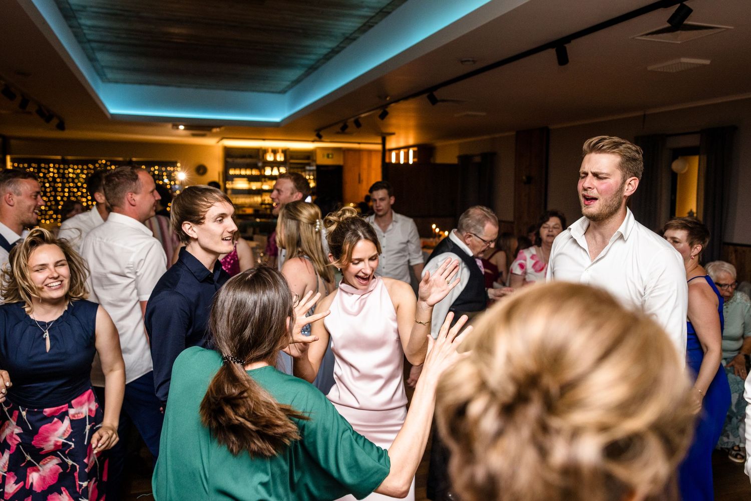 A group of people dancing and socializing at a lively indoor party with blue lighting on the ceiling.