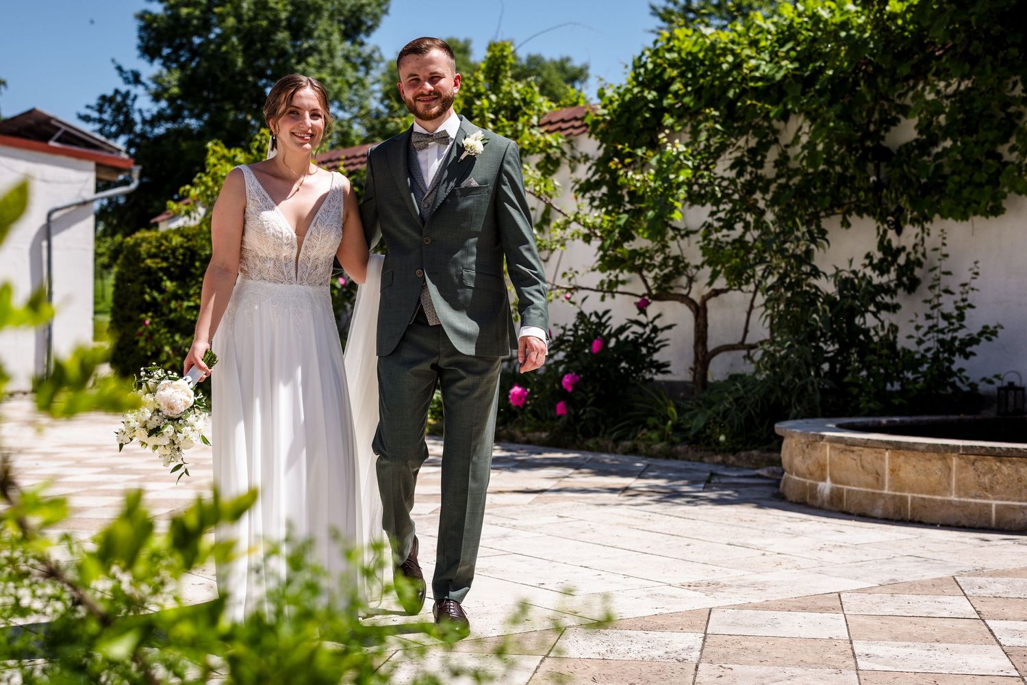 A couple in wedding attire walks together on a sunlit stone patio surrounded by lush greenery and flowers.