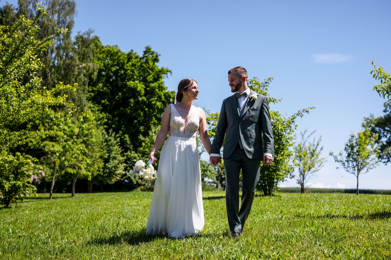 A couple in wedding attire walks together on a lush green hillside under a bright blue summer sky. Fotografiert am Hofgut Sickertshofen