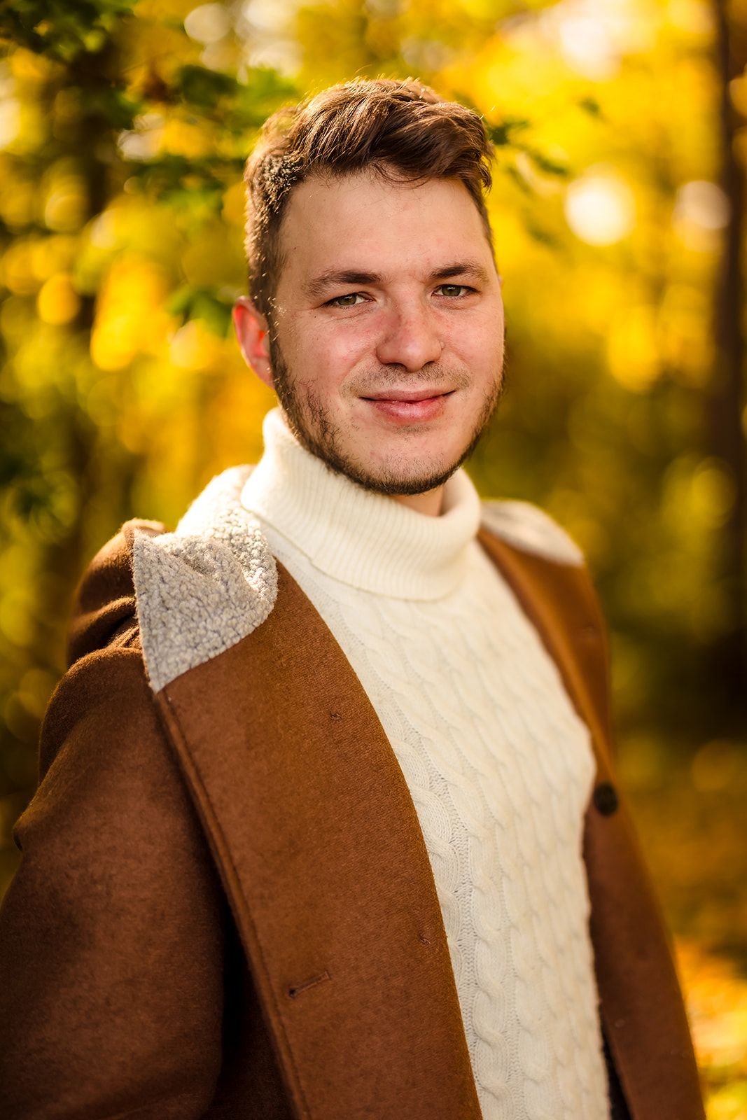 Person in brown jacket and white turtleneck sweater posing in front of golden fall leaves.