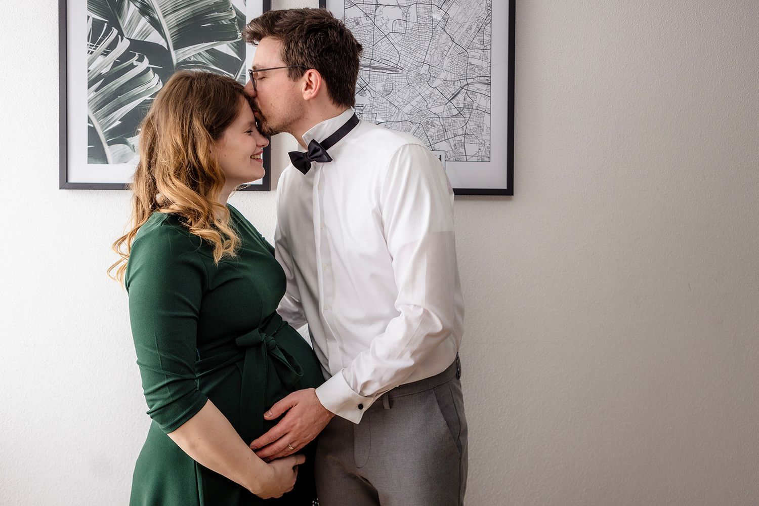 A couple in white and green clothing share an intimate moment against a grey wall with framed artwork.