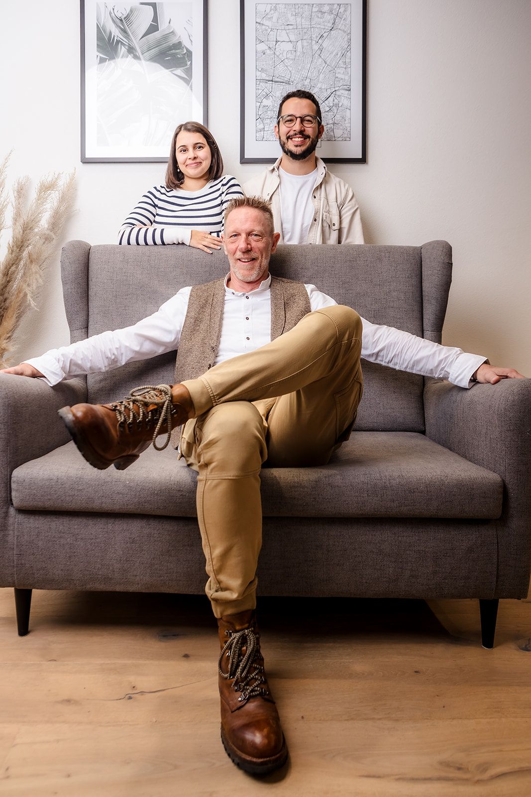 Three people pose together on a modern gray couch in a minimalist living room with framed artwork on the wall.