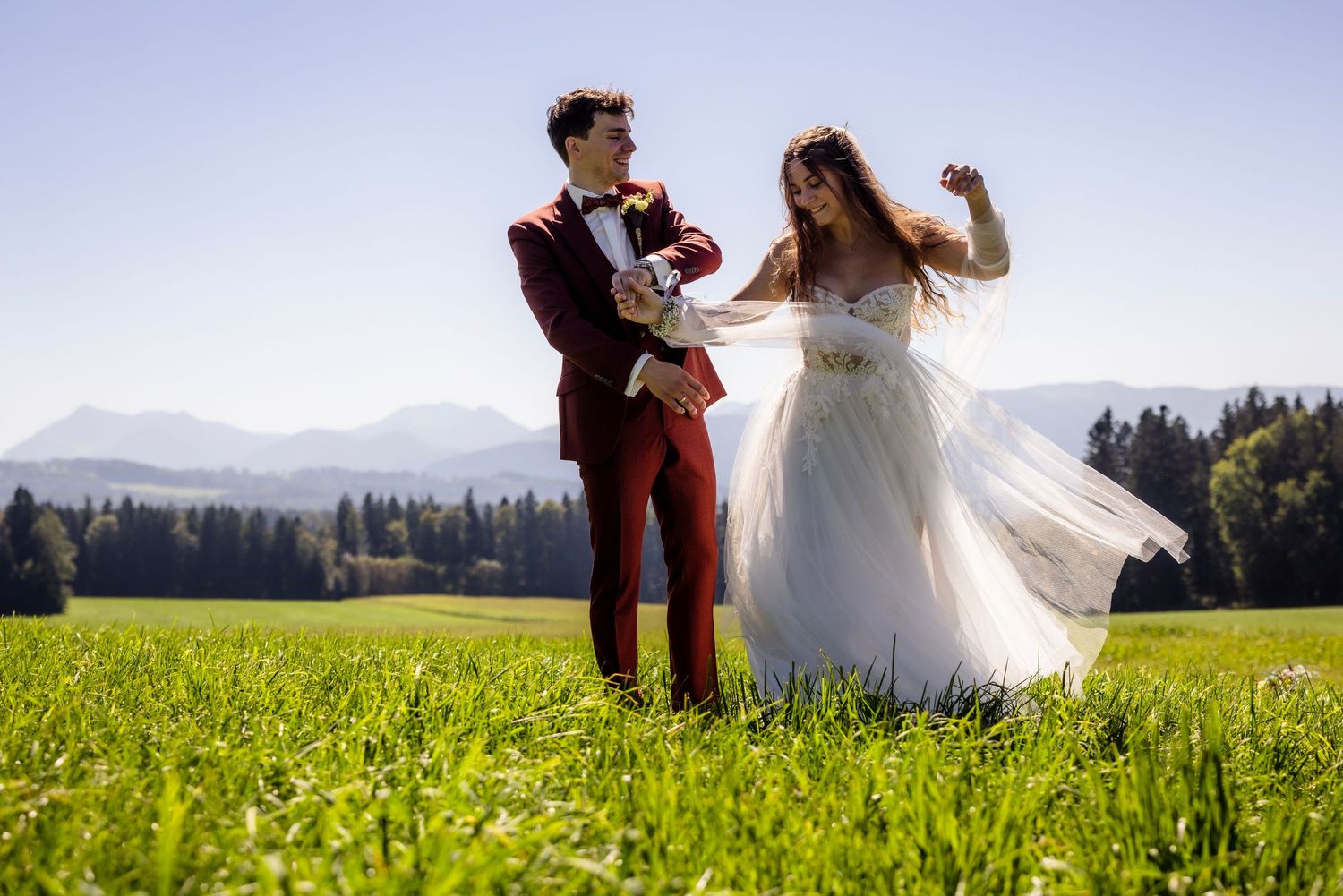 Joyful bride and groom dancing in green meadow with mountain backdrop on sunny wedding day.
