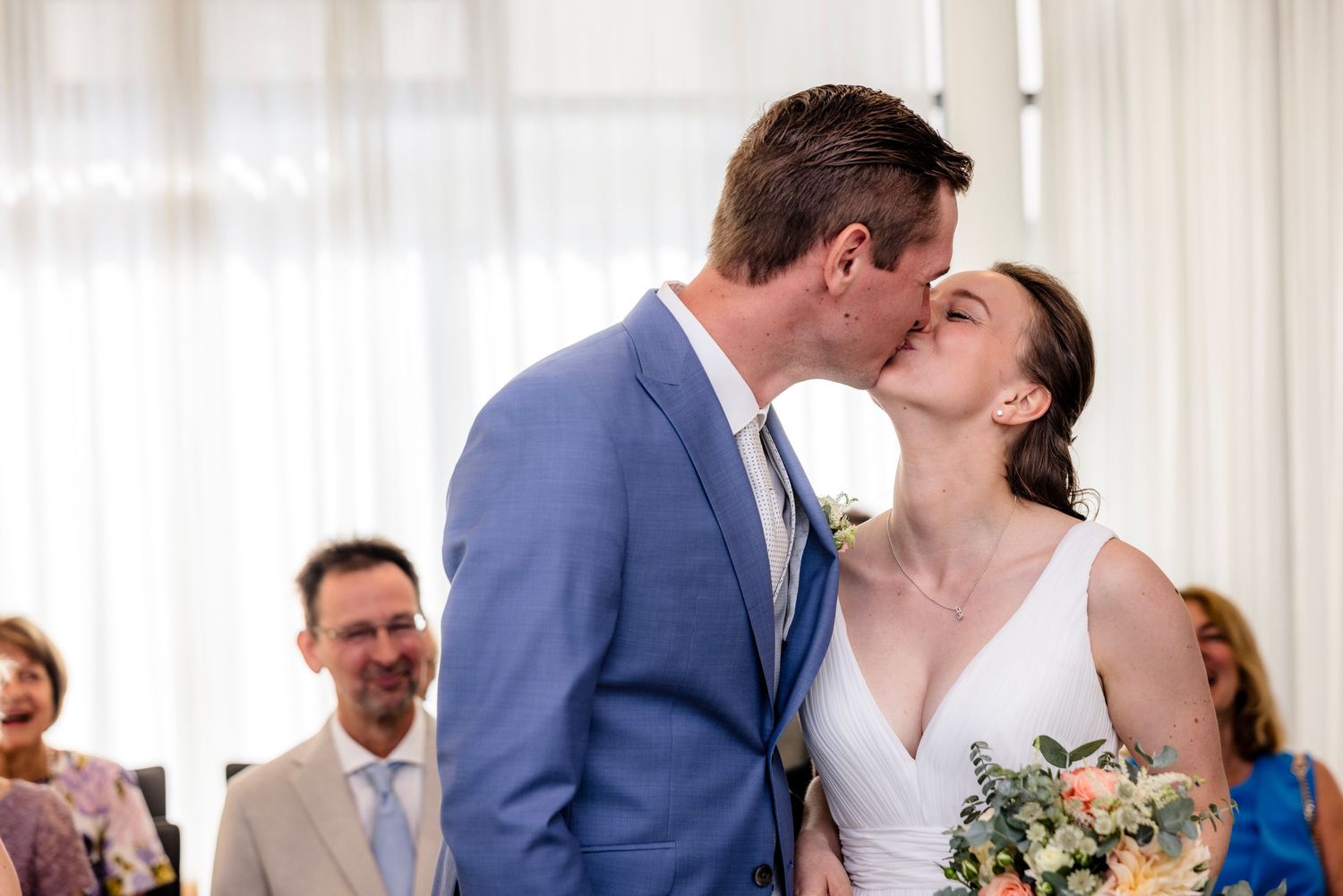 Bride and groom share their first kiss at wedding ceremony while guests smile in background.