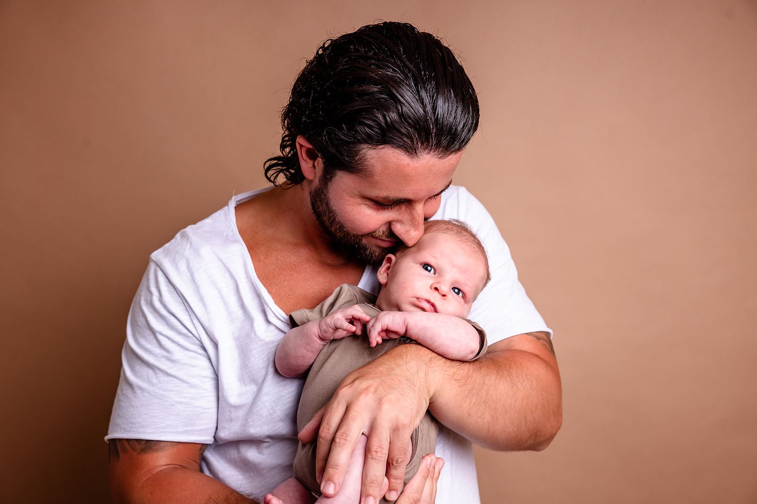 A person wearing a white t-shirt holds and kisses a newborn baby in a tender moment against a warm brown background.
