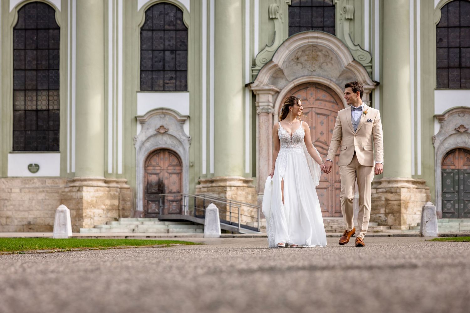 Bride and groom holding hands walking in front of a grand green classical building on their wedding day.