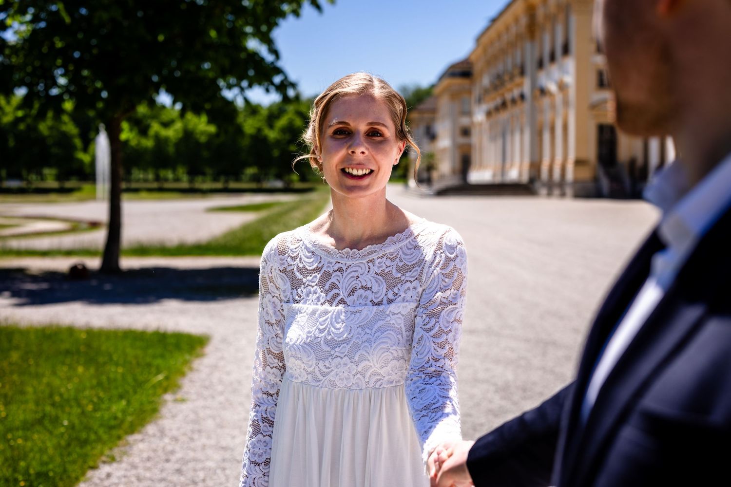 Smiling bride in white lace wedding dress holding hands outdoors near elegant building on sunny day.