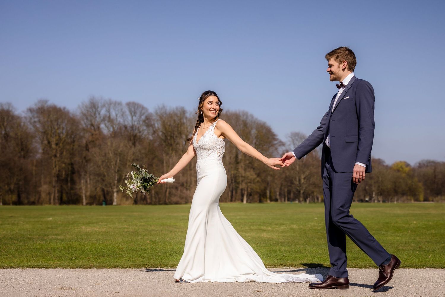Bride and groom walking hand in hand on a sunny day in a lush green park, bride in elegant white gown.