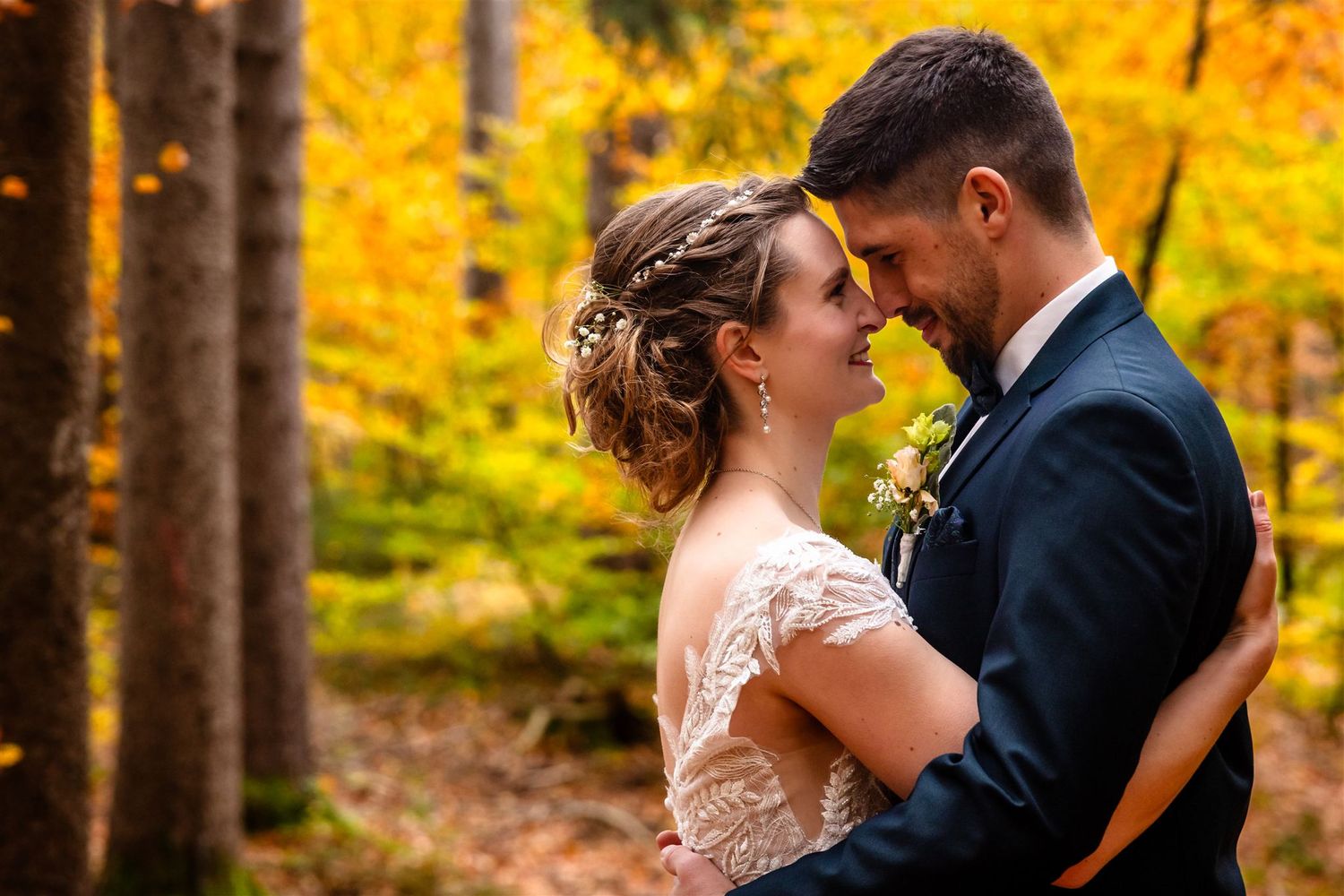 A couple shares a romantic moment in an autumn forest with vibrant yellow foliage in the background.