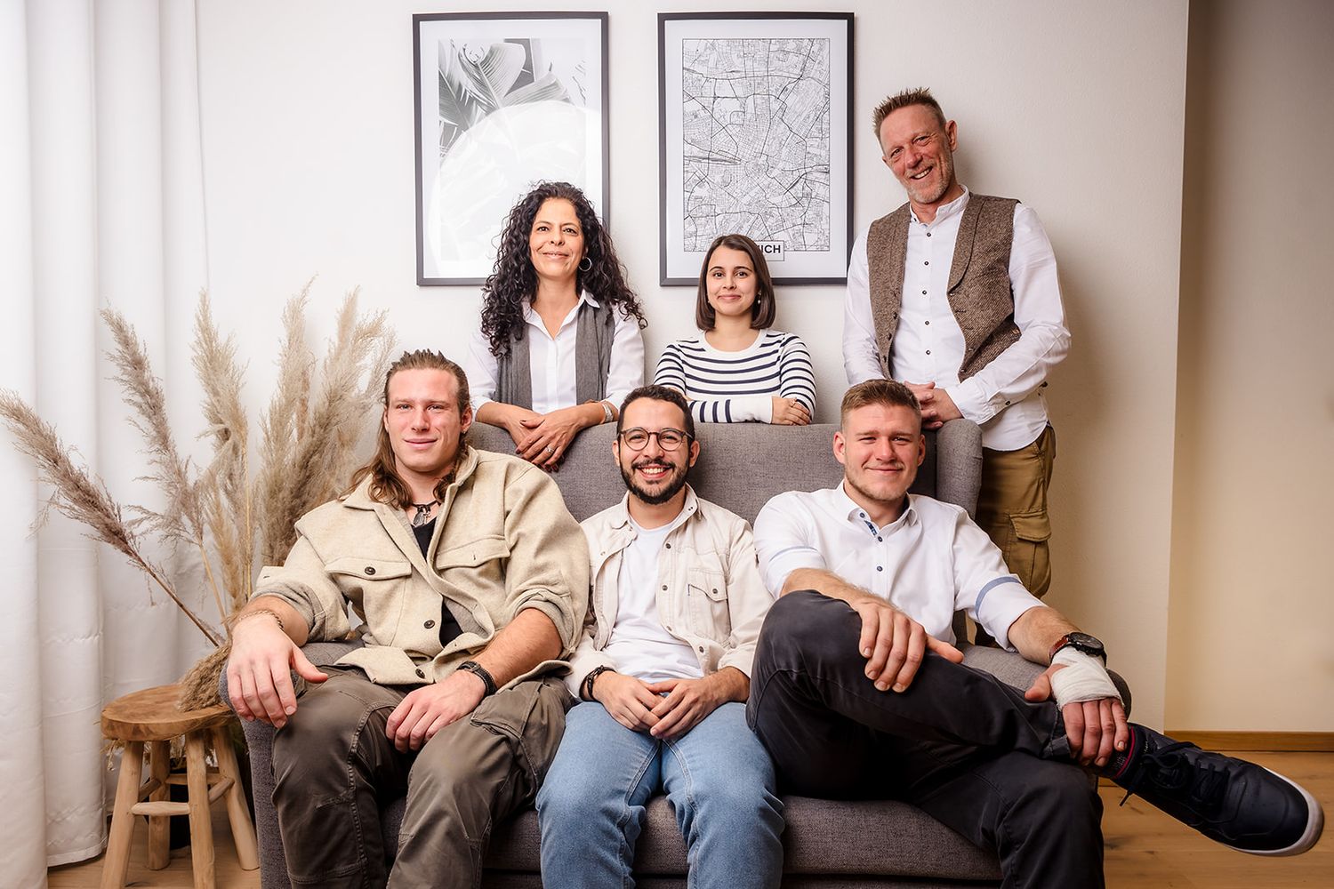 A panoramic group photo of people sitting on a couch in a living room with framed artwork on the wall behind them.