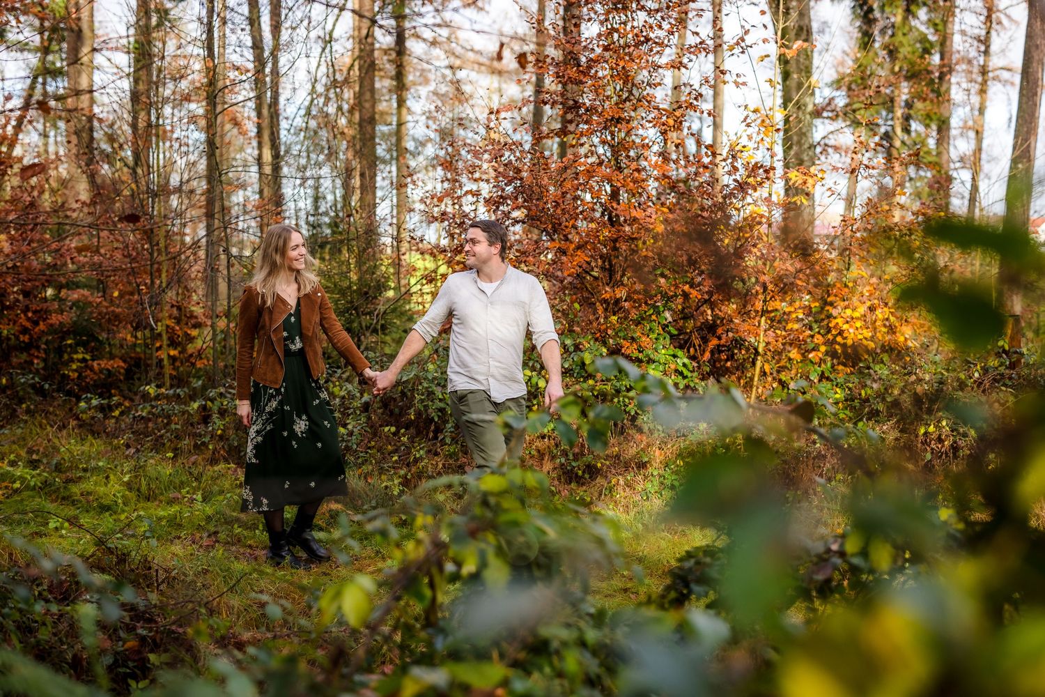 Couple holding hands walking through an autumn forest with golden foliage and warm sunlight filtering through the trees.