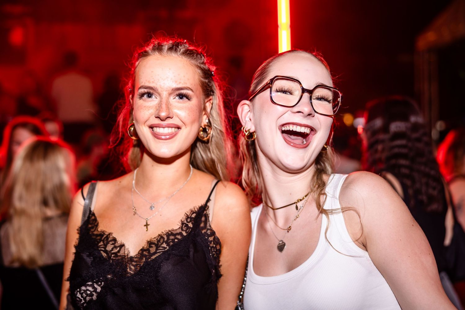 Two smiling young women posing together at a nightclub party with red ambient lighting.