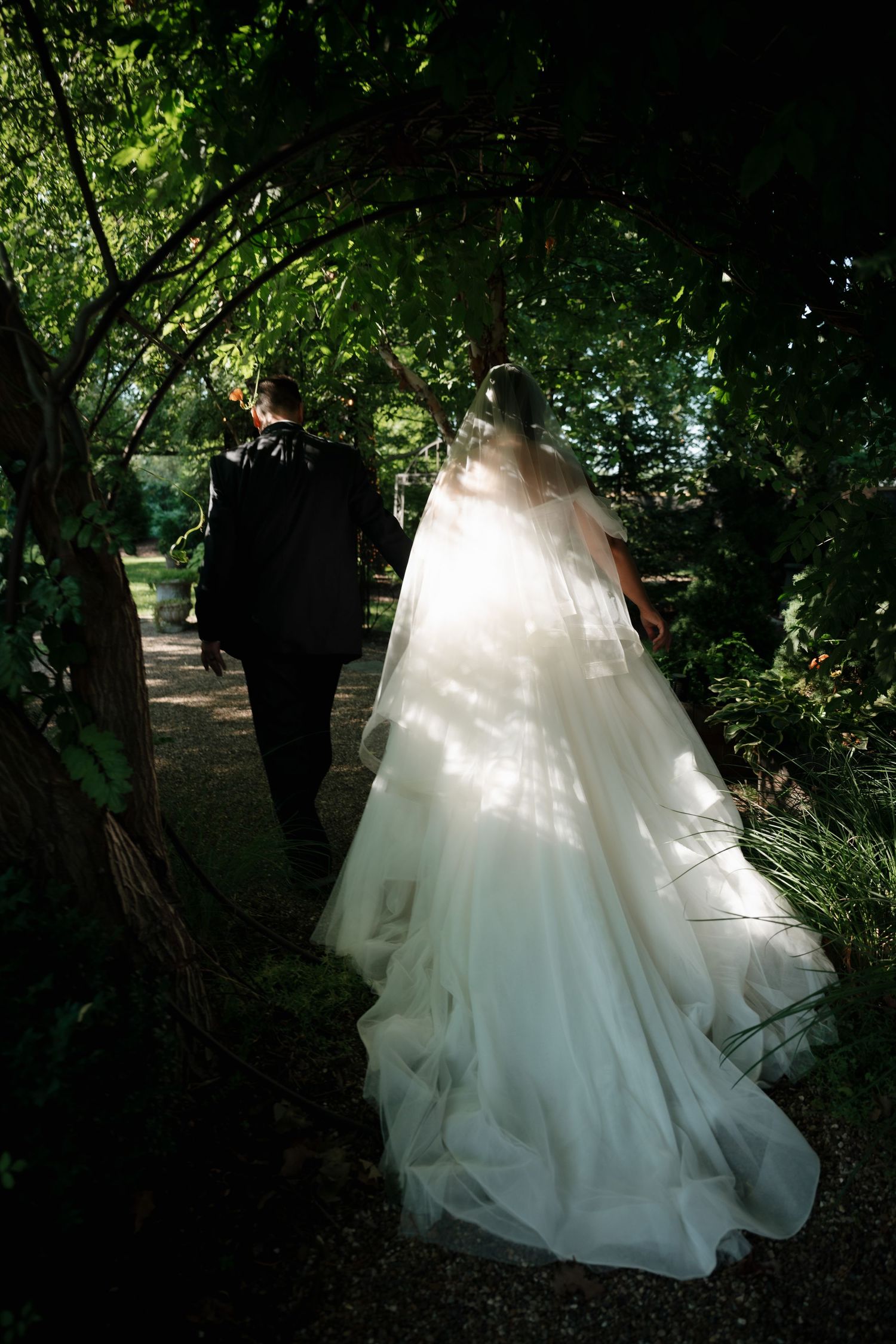 A wedding couple walks through a lush garden path with dappled sunlight filtering through the trees.