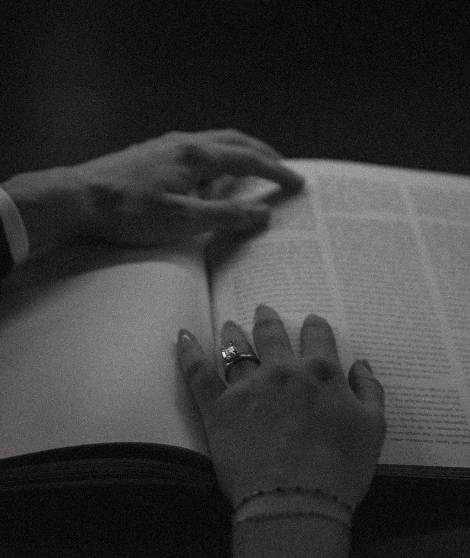 Black and white photo of two hands resting on an open book, one wearing a ring and bracelet.
