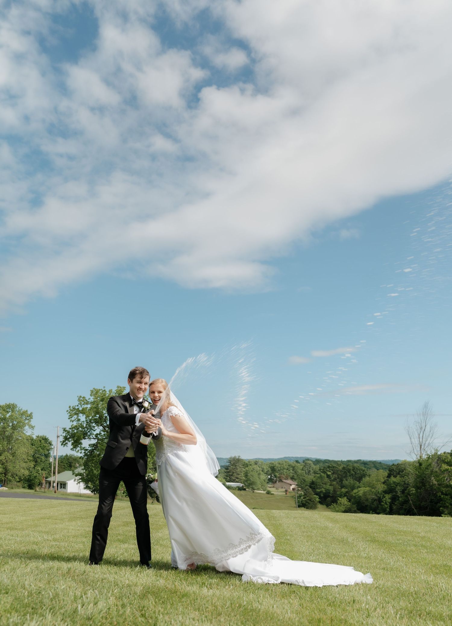 A wedding couple embraces in a sunny meadow with blue skies and white clouds overhead.