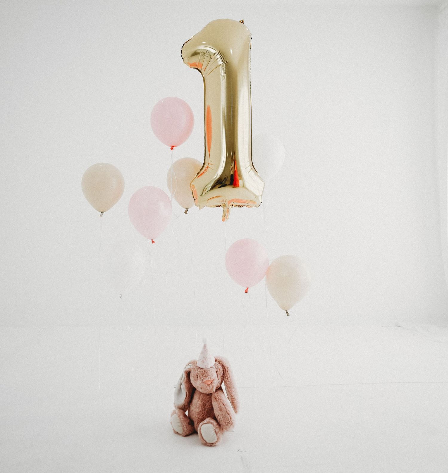 Gold number one balloon with pink and beige balloons floating above a plush bunny toy against white background.