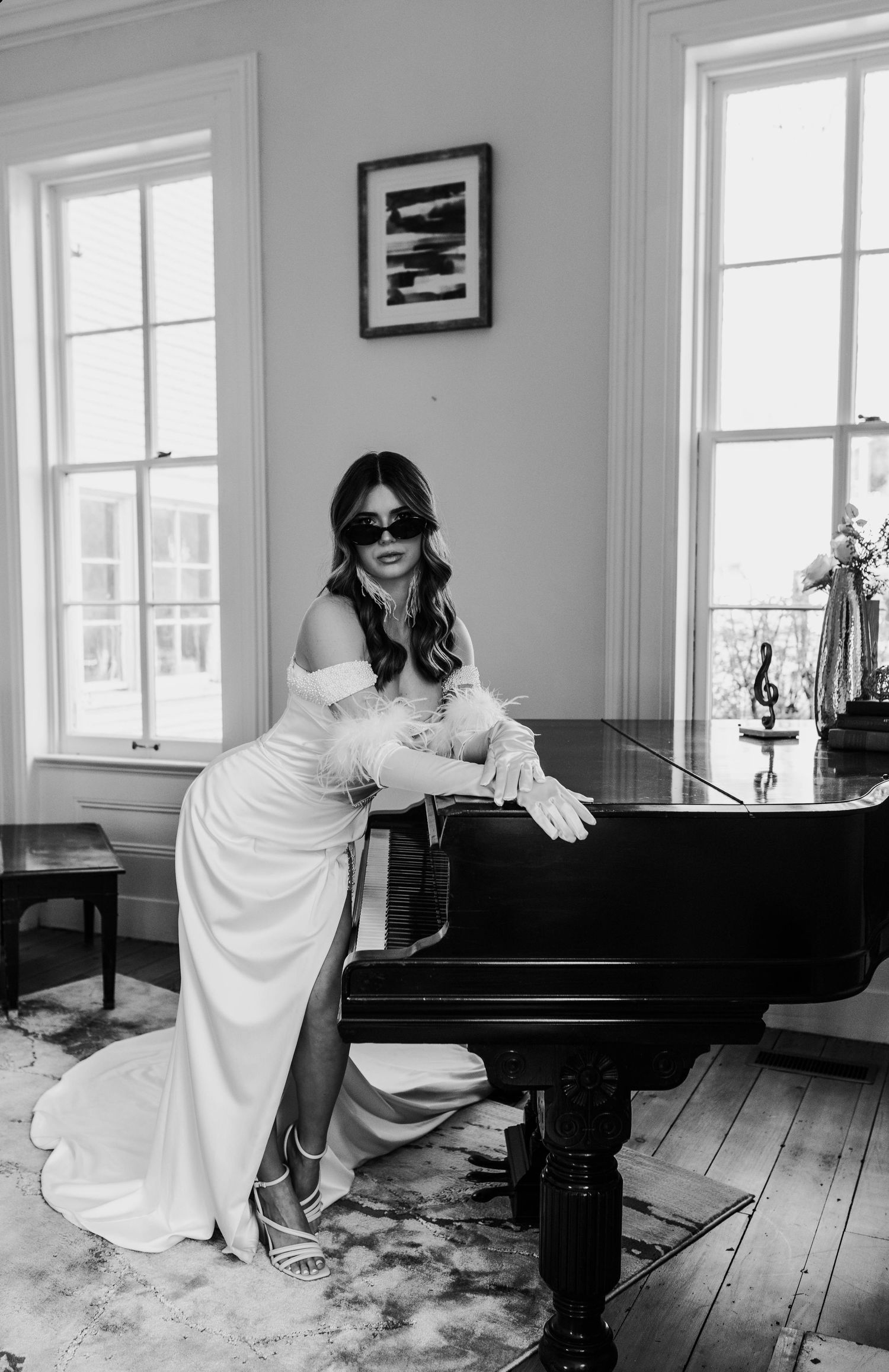 Black and white photo of an elegant figure in a white flowing gown posing at a grand piano in a sunlit room.