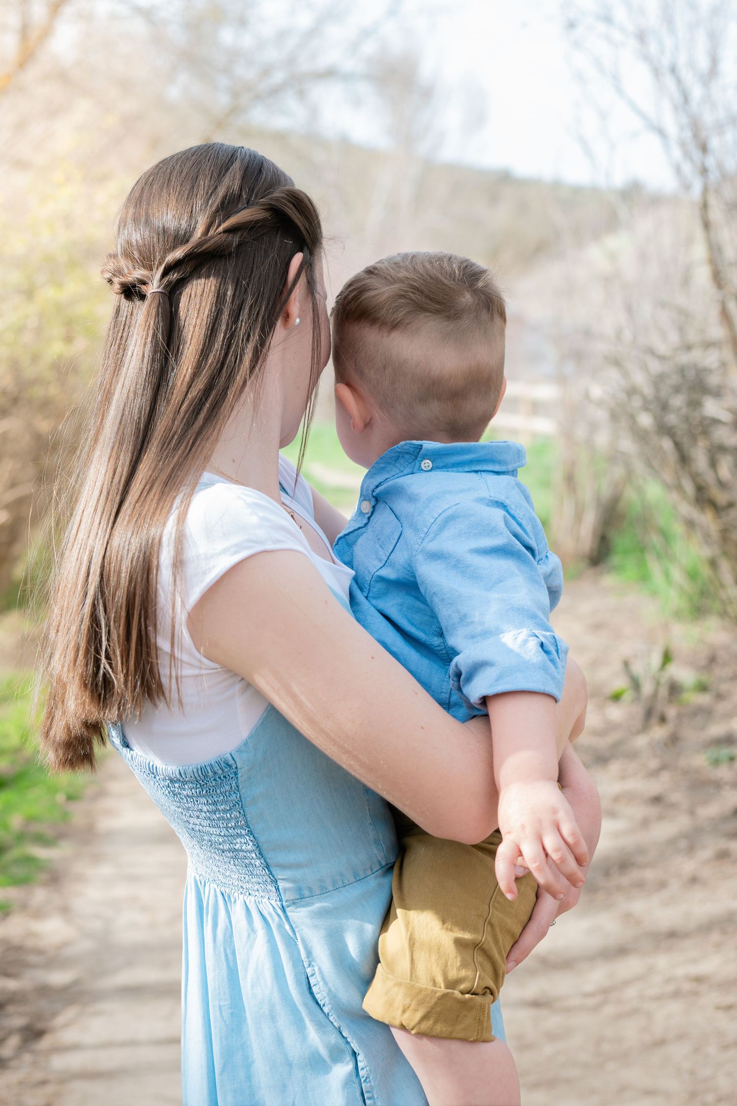 A young mother with long brown hair holds her toddler son outdoors on a sunny path surrounded by trees.
