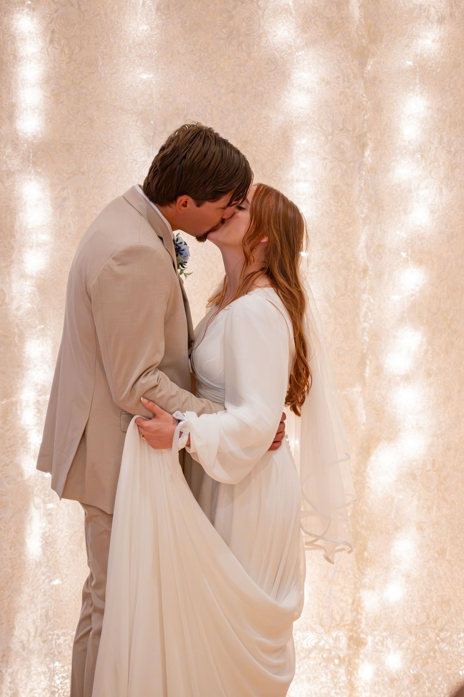 A bride and groom share a romantic first kiss at their wedding, surrounded by a glowing bokeh light backdrop.