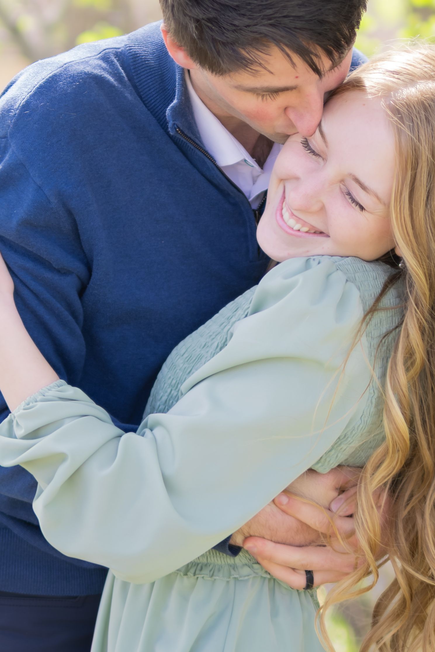 A happy couple embracing outdoors, man in blue sweater kissing smiling blonde woman in mint green jacket.