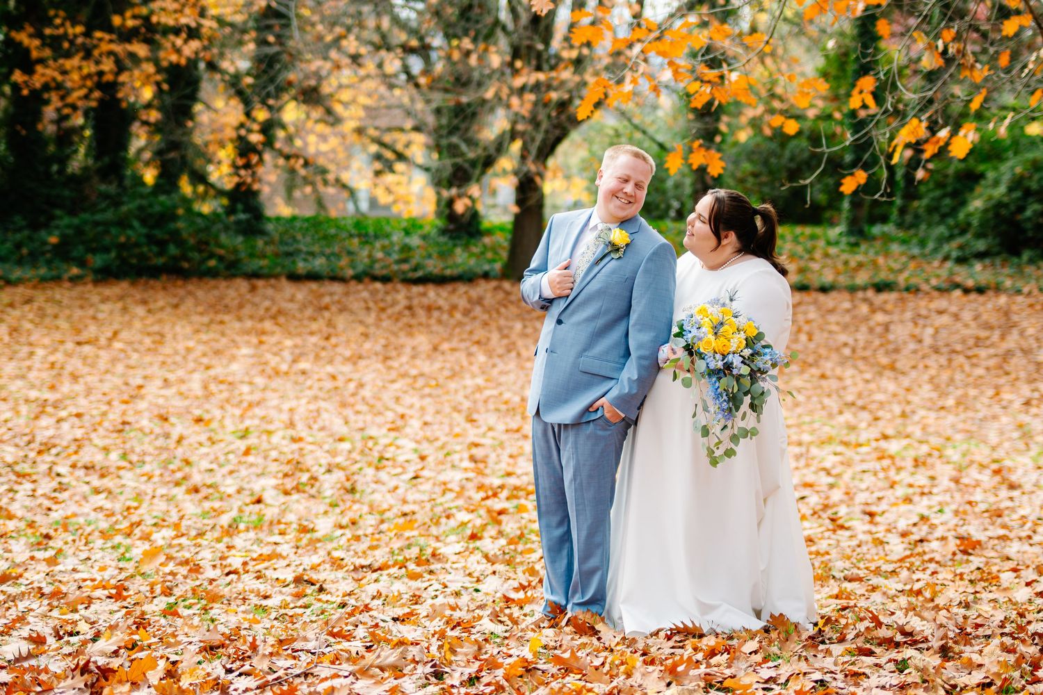 Bride and groom standing on autumn leaves under colorful fall trees, bride holding yellow and blue bouquet.