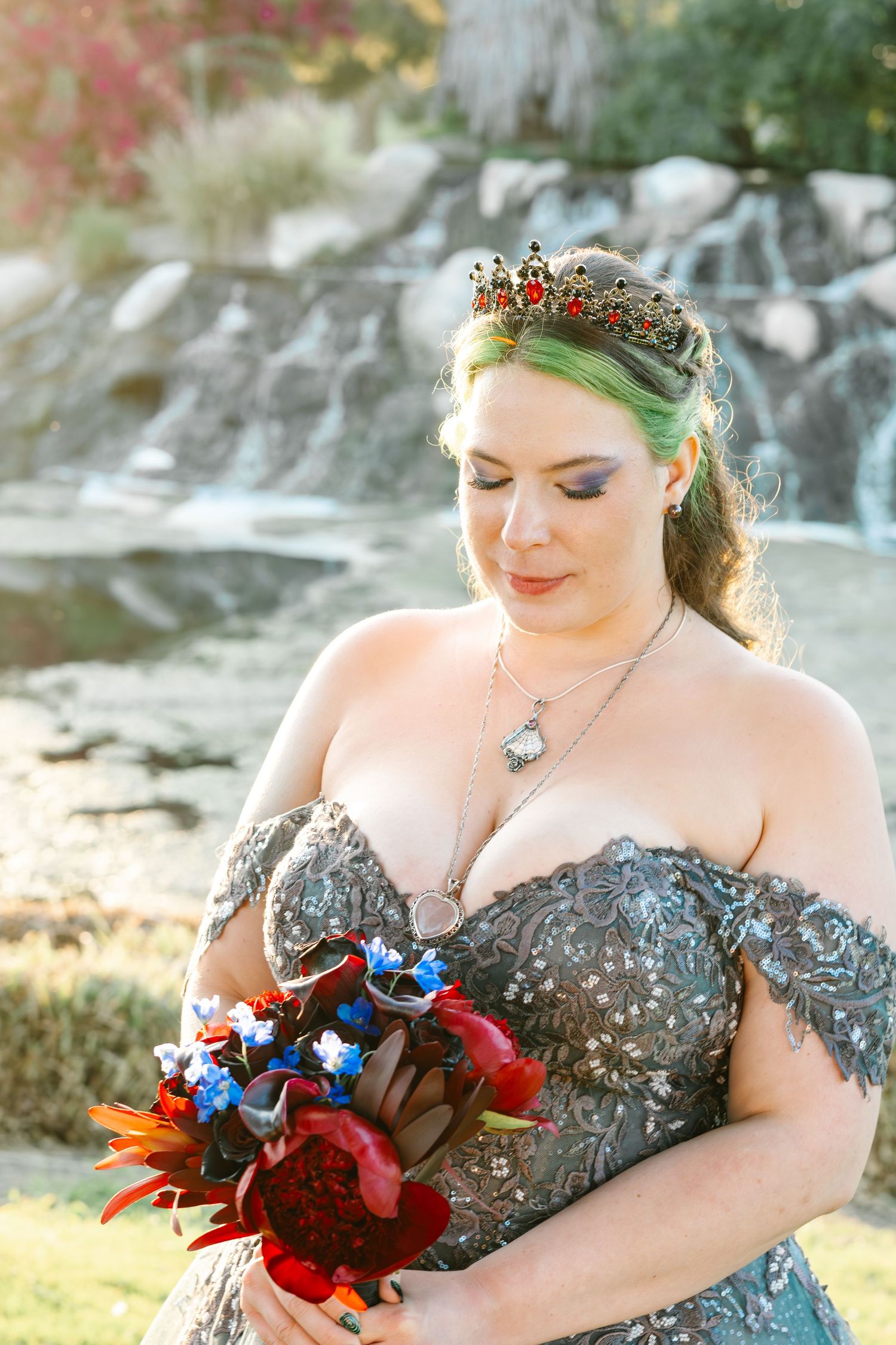 Bride in gray beaded off-shoulder gown with crown holds red and blue bouquet near waterfall.