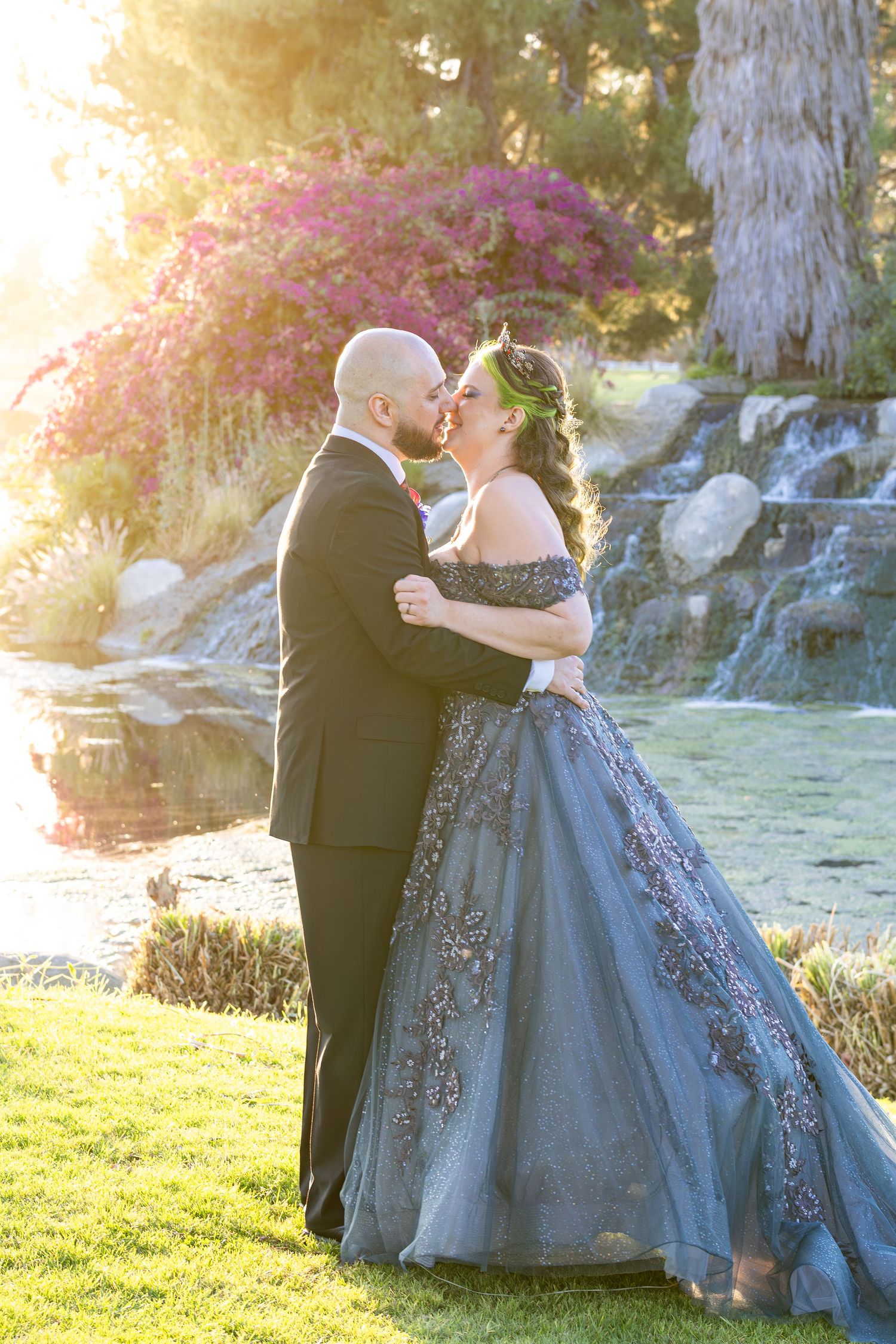 Couple sharing a romantic kiss in a garden, bride in a stunning blue ballgown with floral embroidery, groom in a black tuxedo.