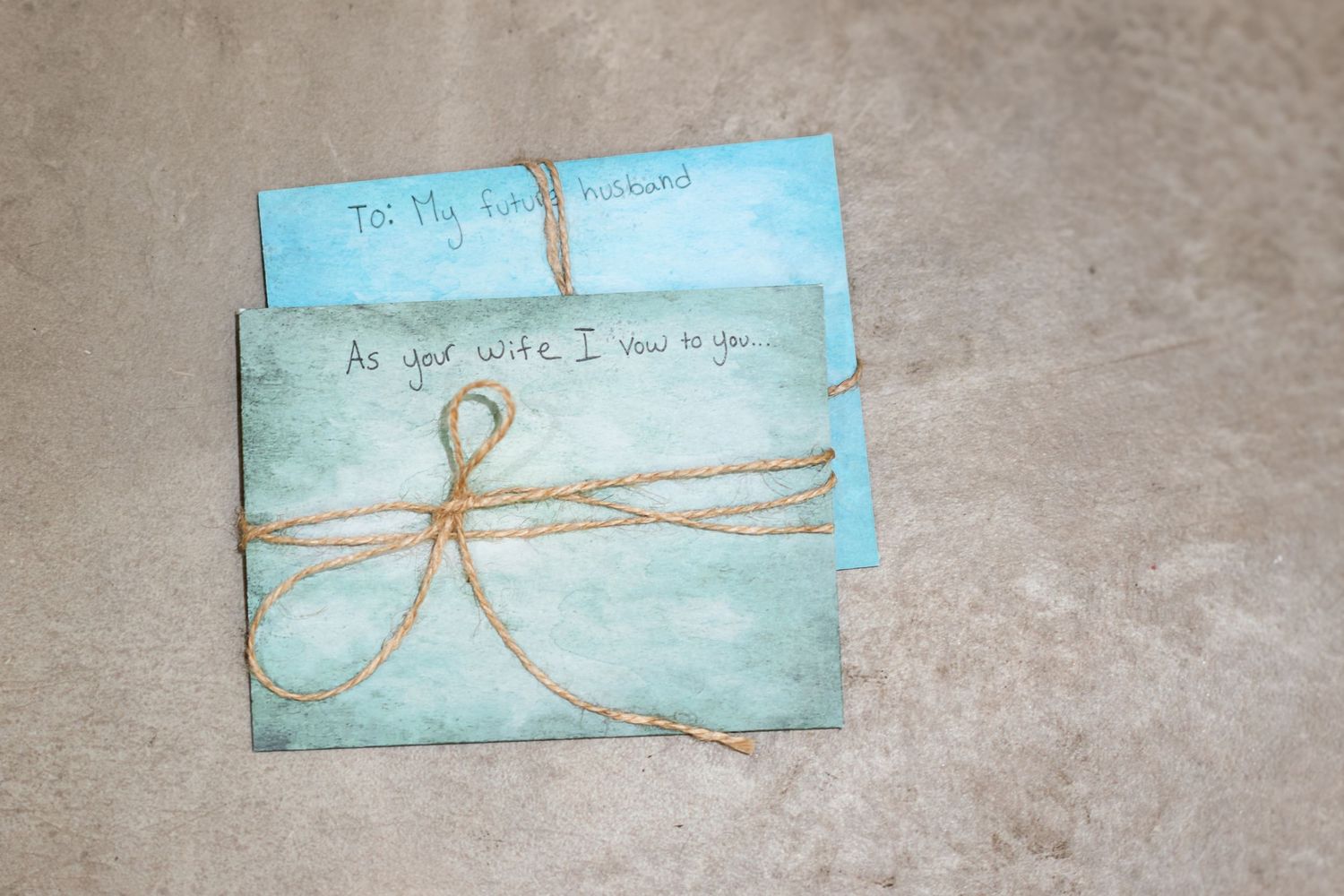 Two light blue wedding vow cards tied with rustic twine, addressed to husband and wife, on a stone surface.