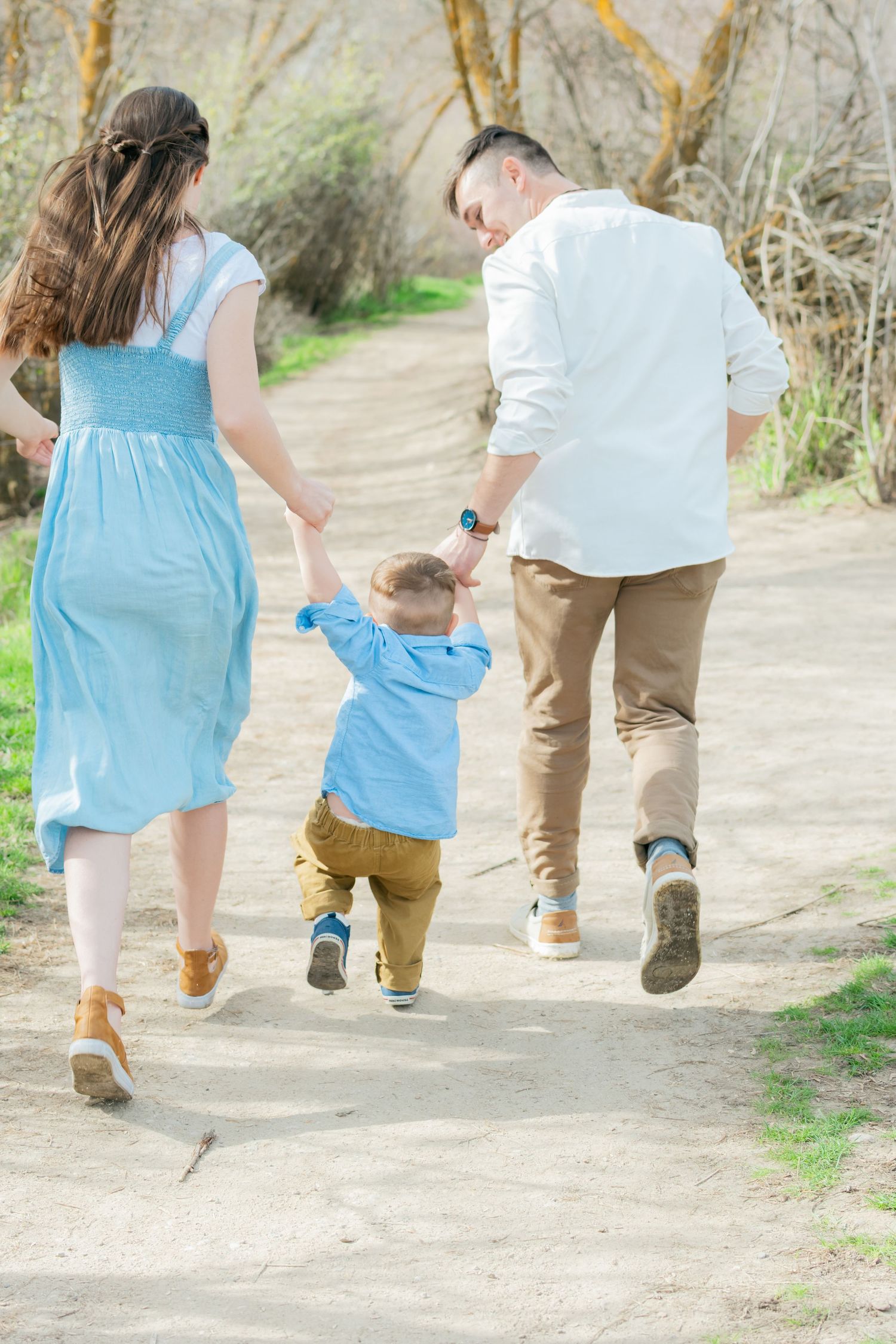 Family of three walking hand-in-hand on a sunny path, parents swinging toddler boy between them outdoors.