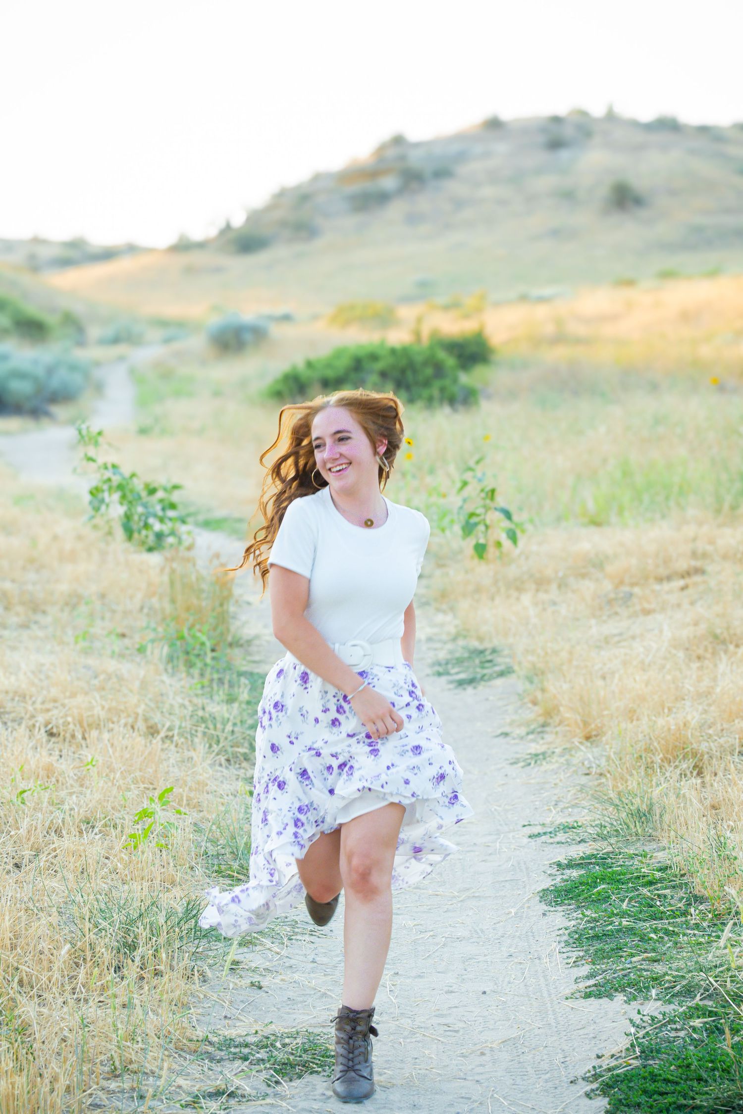 A person in a white top and floral skirt runs playfully on a dirt path through golden grass.
