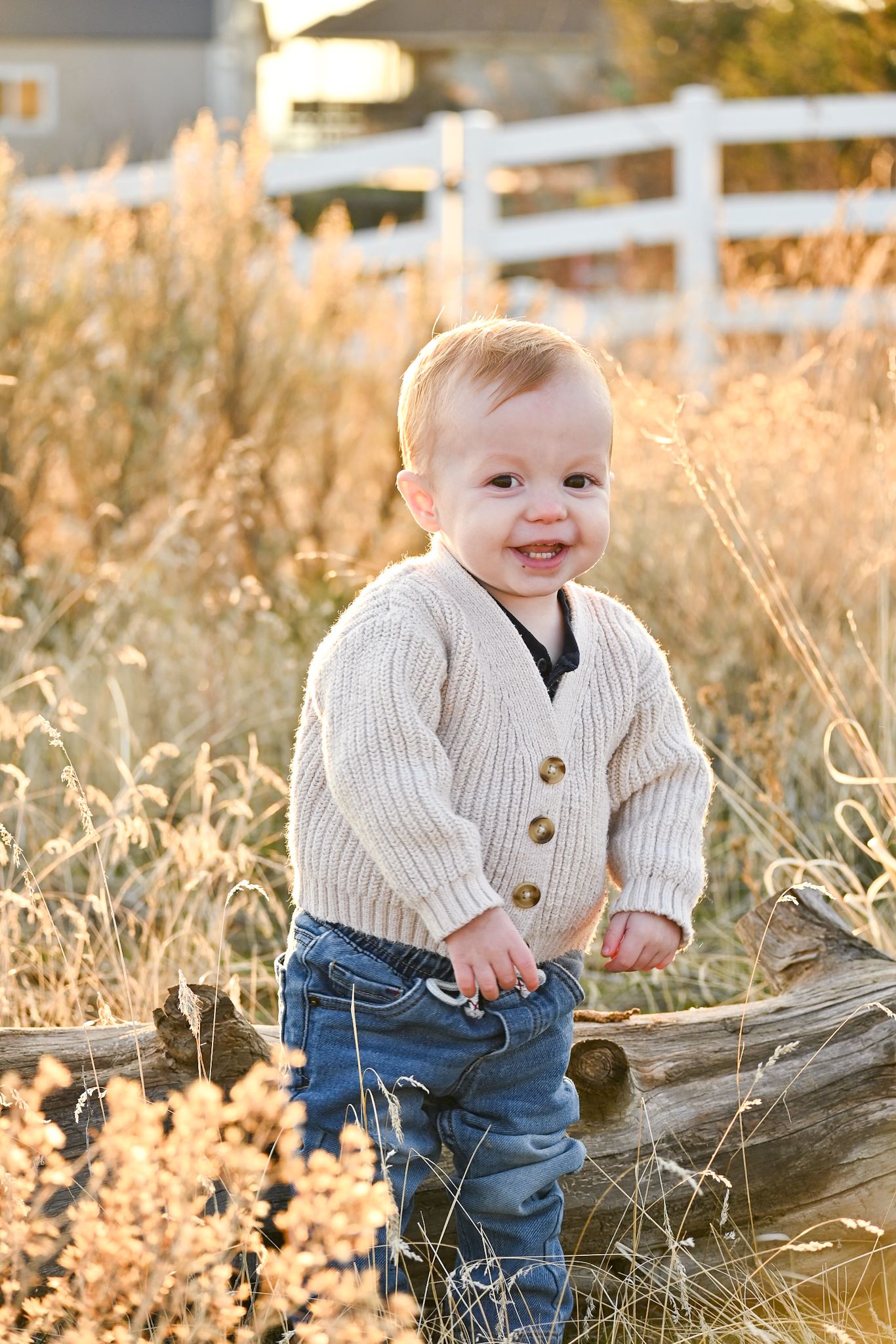 A baby in a white cardigan and jeans sits on a wooden log in golden sunlit grass.
