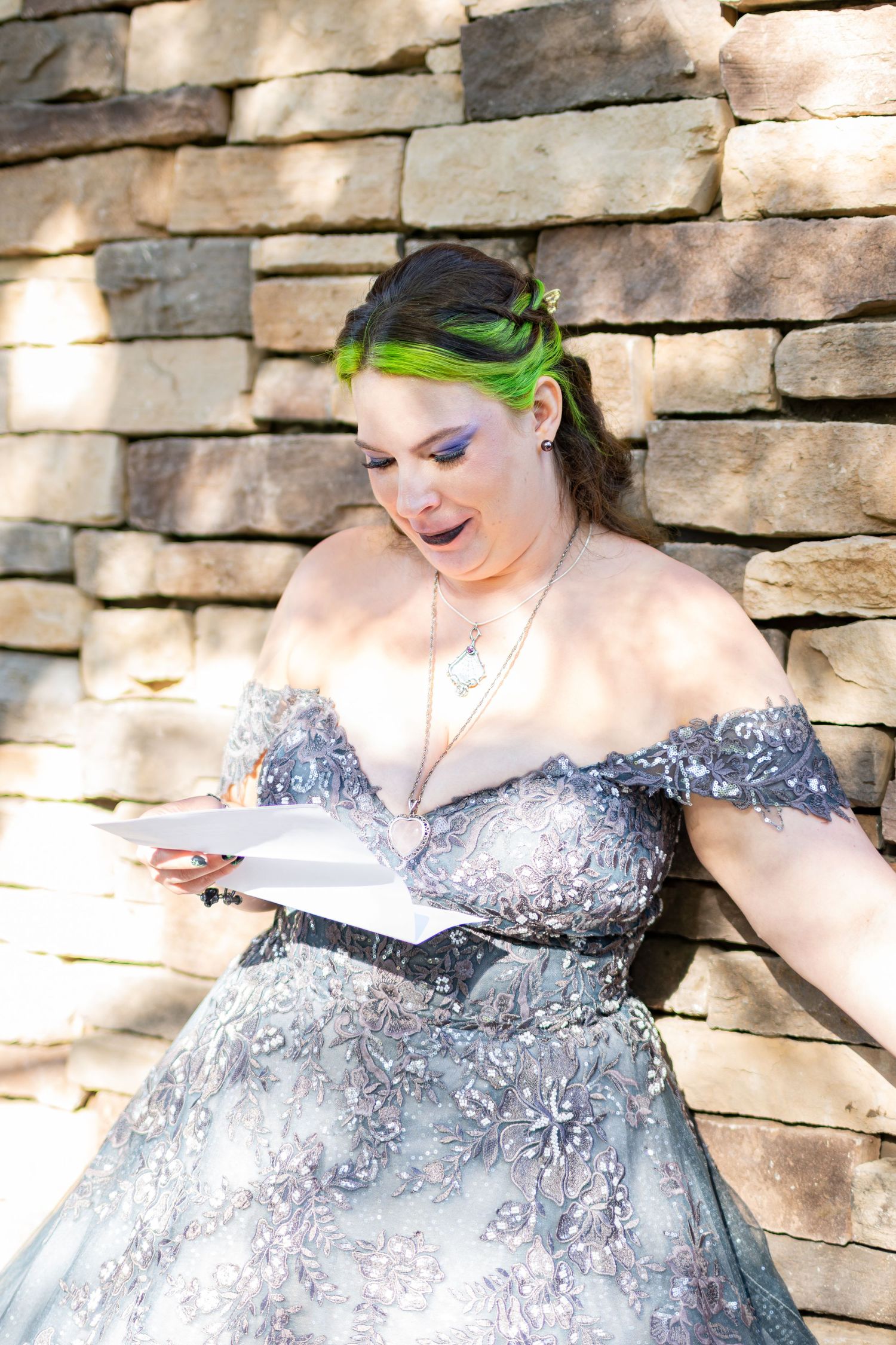 Woman in gray off-shoulder gown with green hair reading vows against stone wall.