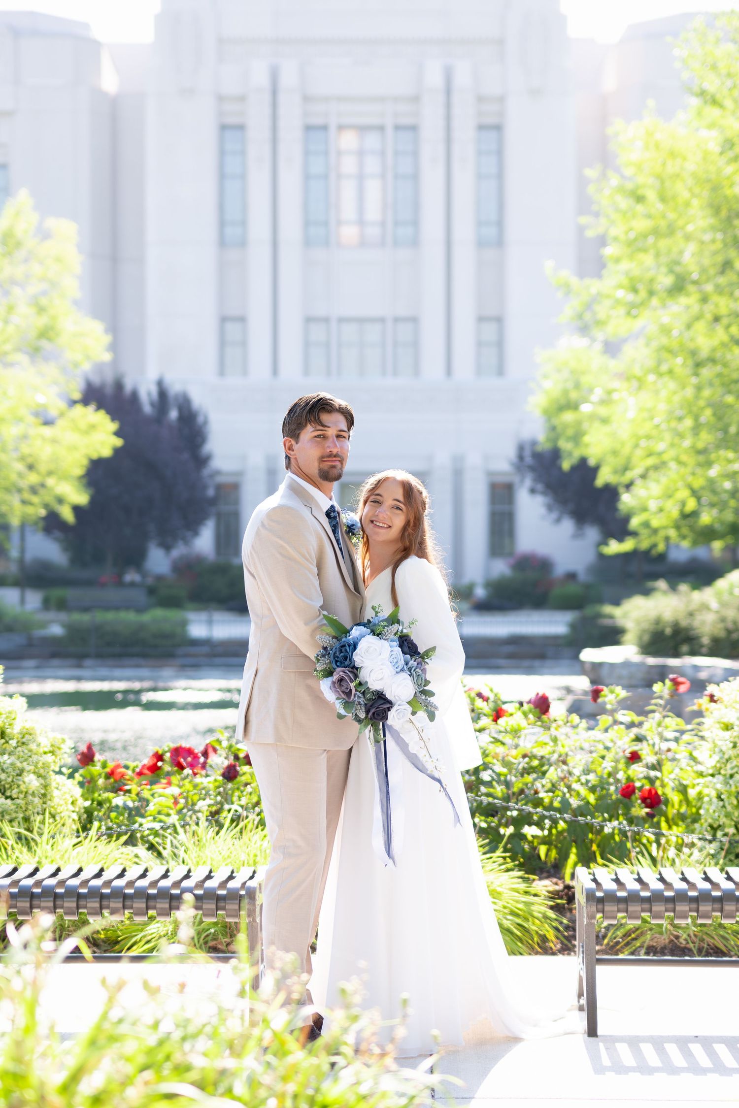 Bride and groom pose in front of a grand white building surrounded by lush gardens on their wedding day.